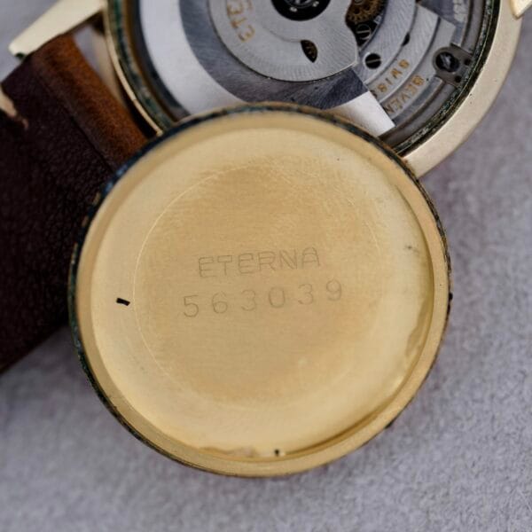 A close-up of the inside back cover of a 1950s Eterna Centaire Eterna Matic Cal. 1428U, showing engraved text ETERNA and number 563039, with part of the movement and a brown leather strap visible.