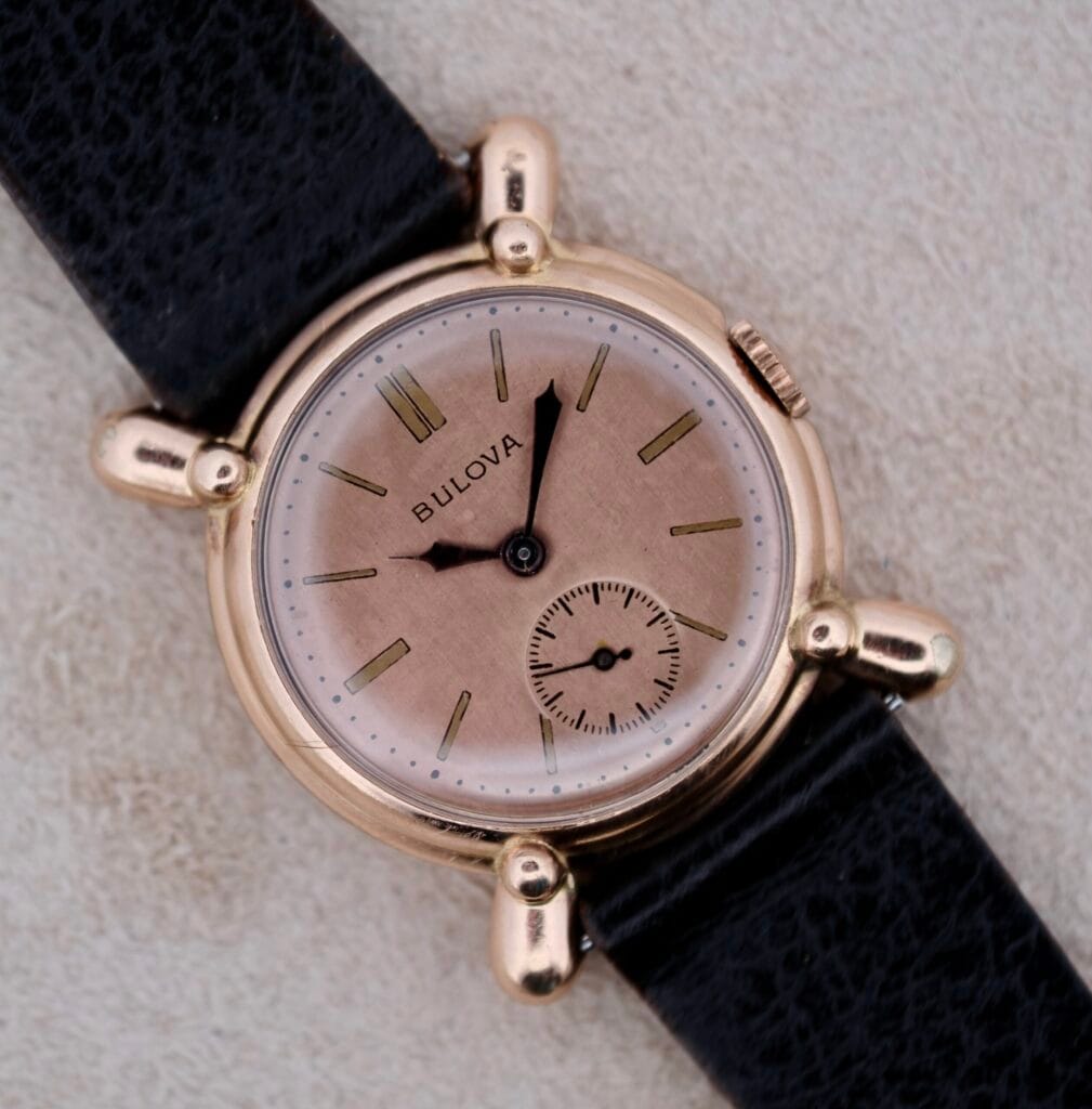A 1940s Bulova Commodore X Shaped Lugs wristwatch features a gold case, pink dial, and black leather strap, displayed on a beige surface.