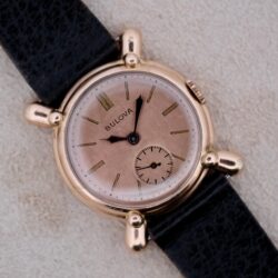 A 1940s Bulova Commodore X Shaped Lugs wristwatch features a gold case, pink dial, and black leather strap, displayed on a beige surface.