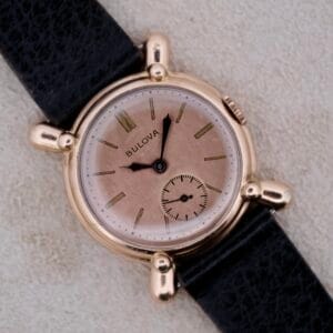 A vintage 1940s Bulova Commodore wristwatch with X-shaped lugs is shown, featuring a round gold-tone case, black leather strap, gold markers, and small seconds subdial at 6 oclock on a light textured background.