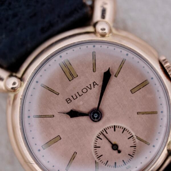 A close-up of a 1940s Bulova Commodore wristwatch with X-shaped lugs, a leather strap, gold-toned case, Roman numerals at 12, 3, 6, and 9, and a small subdial above the 6 o’clock position.