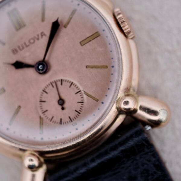 Close-up of a 1940s Bulova Commodore wristwatch featuring X shaped lugs, a round gold-tone case, beige face with small seconds subdial, black leather strap, and visible hands and hour markers.
