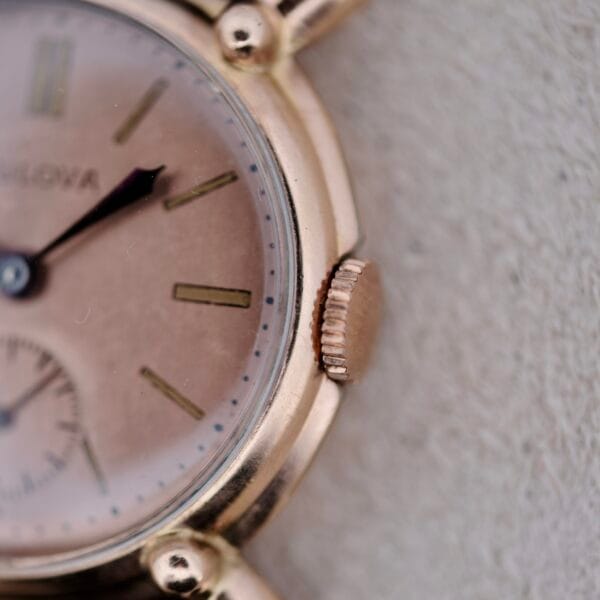 Close-up of a 1940s Bulova Commodore wristwatch face with X-shaped lugs, showing part of the dial, hands, crown, and a partially visible brand name against a light textured background.