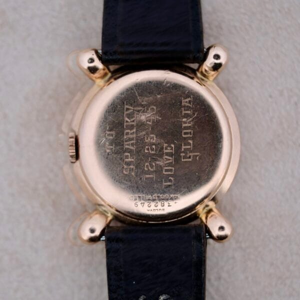 A close-up of the back of a 1940s Bulova Commodore with X-shaped lugs, featuring a black leather strap and an engraving: TO SPARKY 12-25-46 LOVE GLORIA, plus serial numbers and text near the bottom edge on a light grey background.