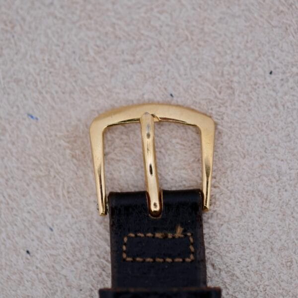 A close-up view of the 1940s Bulova Commodore X Shaped Lugs, featuring a gold-toned metal buckle on a black leather strap with visible stitching, set against a light textured background.