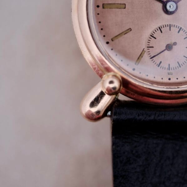 A close-up of a 1940s Bulova Commodore wristwatch shows its gold X-shaped lugs, black leather strap, partial hour markers, gold hands, and small subdial. The face is in focus against a blurred background.