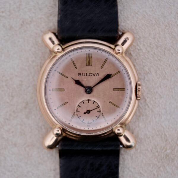 A 1940’s Bulova Commodore wristwatch with X-shaped lugs, a round gold-tone case, black leather strap, beige dial with gold hour markers, black hands, and a small seconds subdial at 6 o’clock.