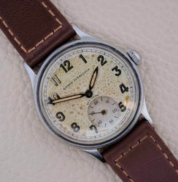 A Girard Perregaux Sea Hawk Military Circa 1943 wristwatch with a brown leather strap, off-white patinated dial, black Arabic numerals, and small seconds at 6 o’clock, photographed on a light textured surface.