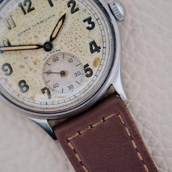 Close-up of a vintage Girard Perregaux Sea Hawk Military Circa 1943 wristwatch featuring a weathered cream dial, black numerals, small seconds subdial, and brown leather strap on a textured light surface.