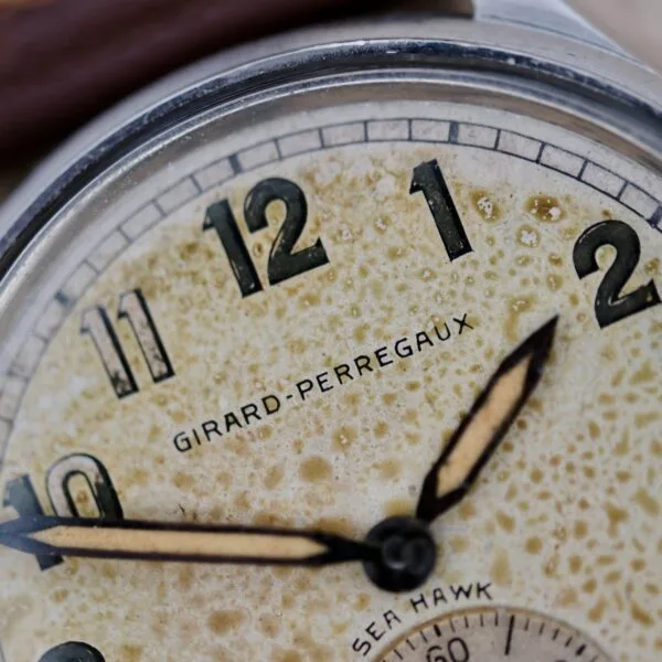 A close-up of a vintage Girard Perregaux Sea Hawk Military Circa 1943 watch face shows worn numerals, patina on the dial, and black-rimmed beige hands at 10:09 with the brand name visible in the center.