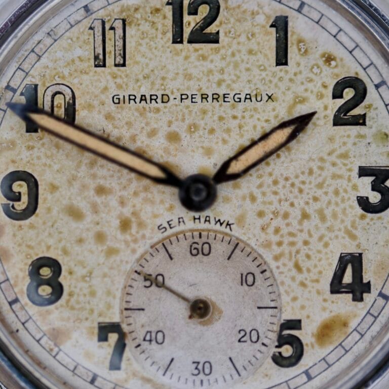 Close-up of an aged Girard Perregaux Sea Hawk Military Circa 1943 watch face with visible discoloration and stains. The time displays about 9:11, and a small seconds subdial appears at the 6 o’clock position.
