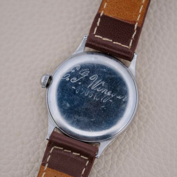 A close-up shows the back of a Girard Perregaux Sea Hawk Military Circa 1943 watch with a brown leather strap and an engraved inscription, E.P. Winger Co. 5403437, on the metal case against a textured beige background.