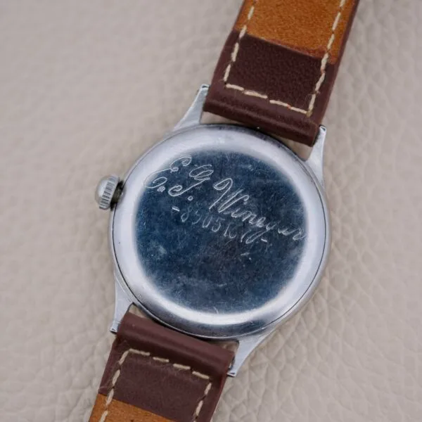 A close-up shows the back of a Girard Perregaux Sea Hawk Military Circa 1943 watch with a brown leather strap and an engraved inscription, E.P. Winger Co. 5403437, on the metal case against a textured beige background.