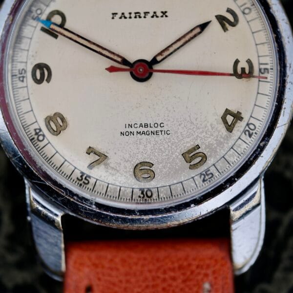 Close-up of a 1940s Fairfax Military Incabloc Manual Wind wristwatch with a worn white dial, gold-tone numerals, silver case, and brown leather strap. The hands show 11:09. Incabloc Non Magnetic is visible on the dial.