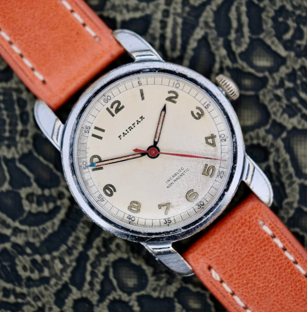 A 1940s Fairfax Military Incabloc Manual Wind wristwatch with a tan dial and orange leather strap, displayed on a lace-patterned background.