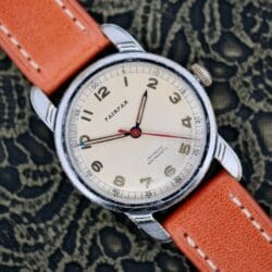 A 1940s Fairfax Military Incabloc Manual Wind wristwatch with a tan dial and orange leather strap, displayed on a lace-patterned background.