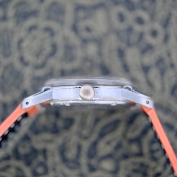 A close-up side view of the 1940s Fairfax Military Incabloc Manual Wind watch with a metallic case and round crown, set against a patterned background. The orange and black strap is partially visible on both sides.