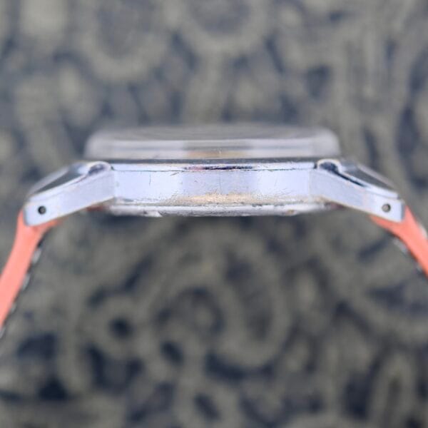 Close-up side view of the 1940s Fairfax Military Incabloc Manual Wind wristwatch, featuring a scratched, worn metal case and visible lugs; background is out of focus with a dark and light patterned design.