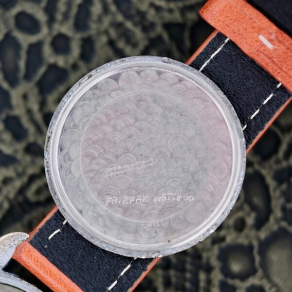 A close-up of the inside case back of a 1940s Fairfax Military Incabloc Manual Wind wristwatch, showing faint engraved markings, displayed on a black and brown leather strap against a textured background.