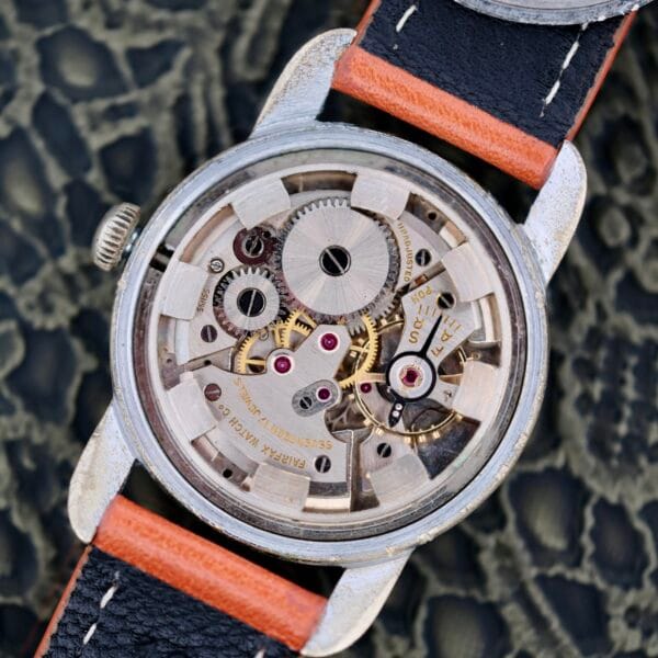 A close-up of the 1940s Fairfax Military Incabloc Manual Wind wristwatch with its case back removed, revealing intricate mechanical gears and movement parts. The watch features a leather strap with orange and black segments.