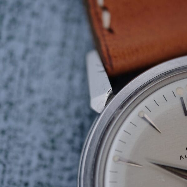 Close-up of the Omega Seamaster Automatic 2832-15, featuring a silver case, brown leather strap, and silver hour markers with a second hand, set against a textured blue-gray background.