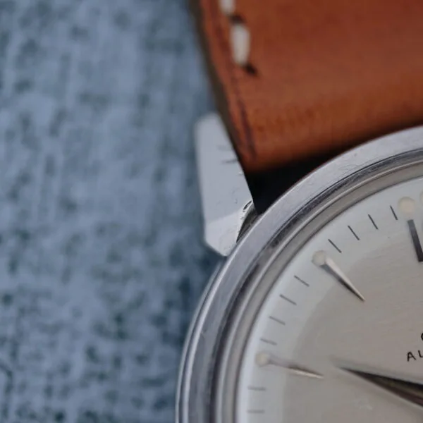 Close-up of the Omega Seamaster Automatic 2832-15, featuring a silver case, brown leather strap, and silver hour markers with a second hand, set against a textured blue-gray background.