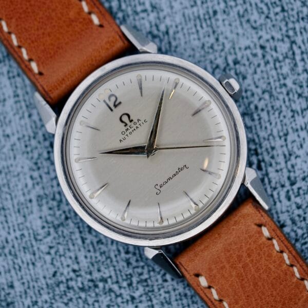 A close-up of the Omega Seamaster Automatic 2832-15 wristwatch, featuring a silver dial, black hour markers, silver hands, and a brown leather strap, displayed on a textured gray surface.