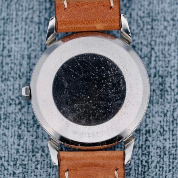 A close-up of the back of an Omega Seamaster Automatic 2832-15 wristwatch with a brown leather strap shows a scratched metal case back, a circular black mark at the center, and the word WATERPROOF faintly visible near the bottom.