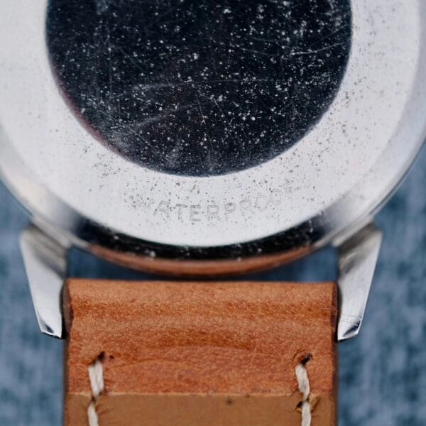 Close-up of the back of an Omega Seamaster Automatic 2832-15 wristwatch with a worn metal case and scratches, attached to a brown leather strap with visible stitching. WATERPROOF is faintly engraved on the case.