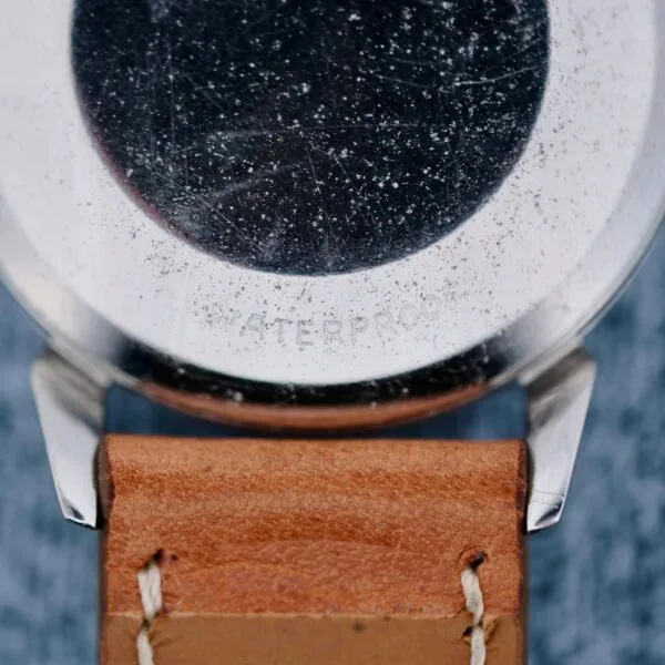 Close-up of the back of an Omega Seamaster Automatic 2832-15 wristwatch with a worn metal case and scratches, attached to a brown leather strap with visible stitching. WATERPROOF is faintly engraved on the case.