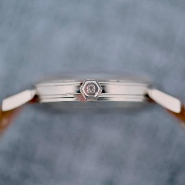 Close-up side view of the Omega Seamaster Automatic 2832-15, a silver wristwatch with a hexagonal crown featuring an engraved logo, set against a blurred gray background.