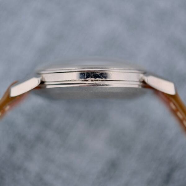 Close-up side view of the Omega Seamaster Automatic 2832-15, featuring a silver thin case with no visible markings, attached to a light brown strap, set against a blurred gray background.