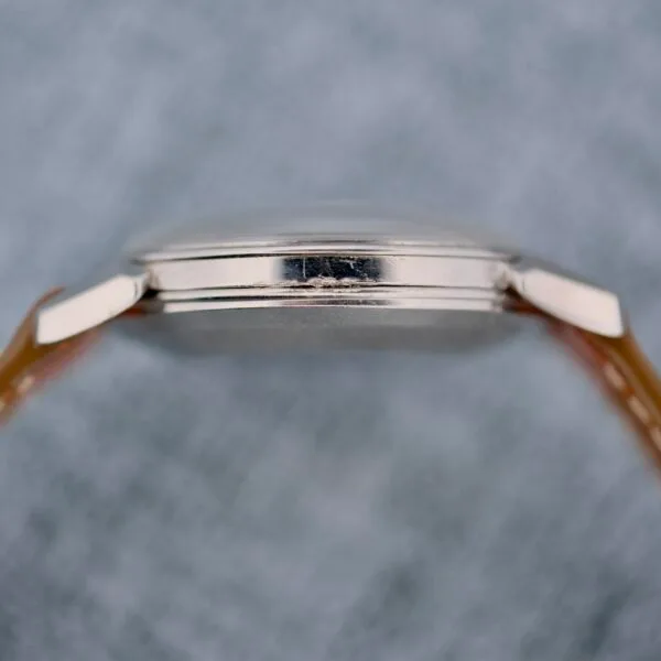 Close-up side view of the Omega Seamaster Automatic 2832-15, featuring a silver thin case with no visible markings, attached to a light brown strap, set against a blurred gray background.