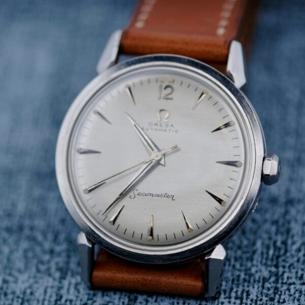 A close-up of an Omega Seamaster Automatic 2832-15 wristwatch with a silver dial, silver hour markers, and a brown leather strap, set against a textured blue background.