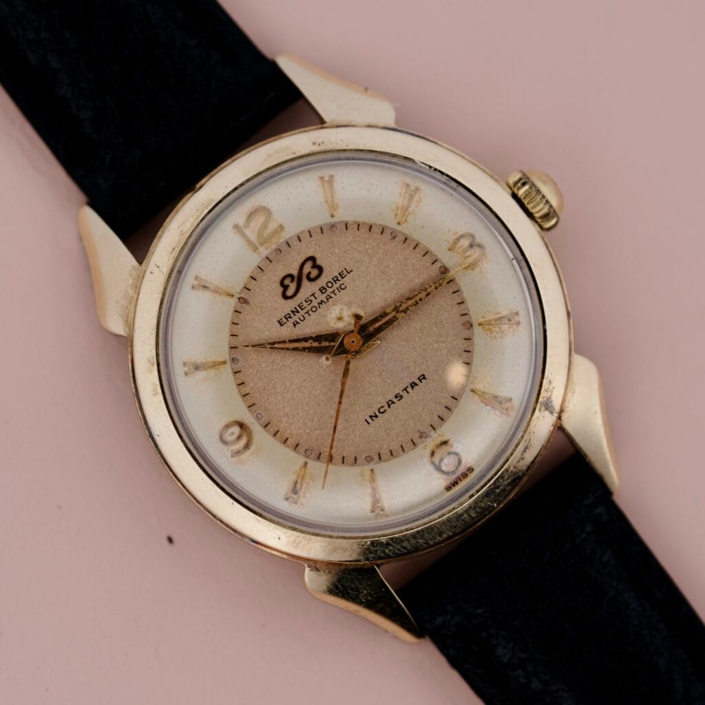 Ernest Borel Incastar 1560 Gold Capped wristwatch with black leather strap and beige dial, displayed on a pink background.