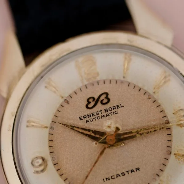 A close-up of an Ernest Borel Incastar 1560 Gold Capped wristwatch highlights the watch face, gold hour markers, black hour hand, and visible wear and scratches on the glass and dial.