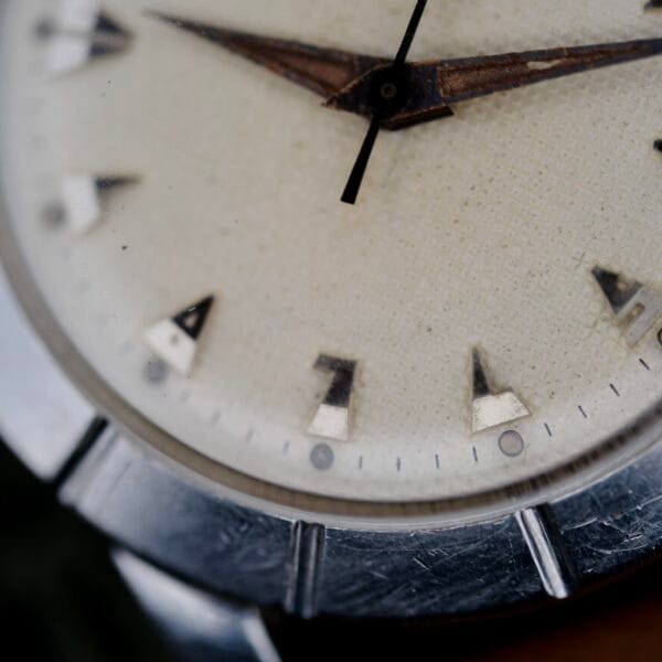 Close-up of the Wittnauer 2065 Waffle Dial wristwatch, featuring an analog display with metallic Art Deco indices and a textured off-white dial, plus a scratched, worn bezel partially visible.