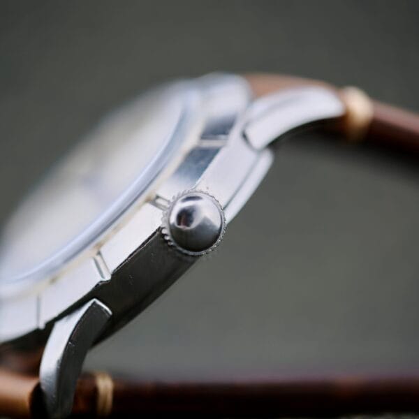 Close-up side view of the Wittnauer 2065 Waffle Dial Art Deco Indicies, focusing on the crown and part of the metal case with a brown leather strap, set against a blurred green background.