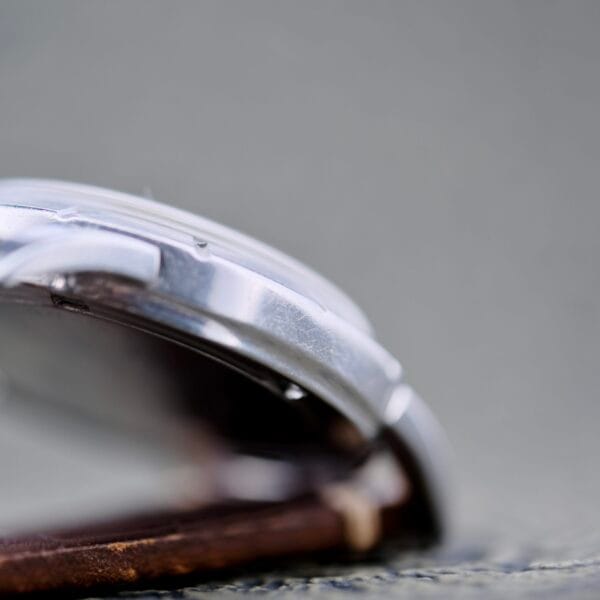 A close-up side view of the Wittnauer 2065 Waffle Dial Art Deco Indicies wristwatch with a brown leather strap, highlighting the silver metal case and crystal against a blurred gray background.
