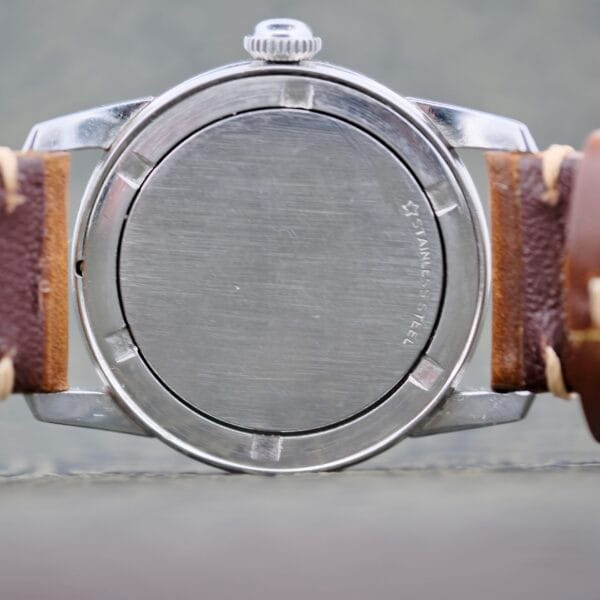 A close-up of the Wittnauer 2065 Waffle Dial Art Deco Indicies shows its stainless steel case, brown leather straps with visible stitching, and the engraving STAINLESS STEEL on the back.