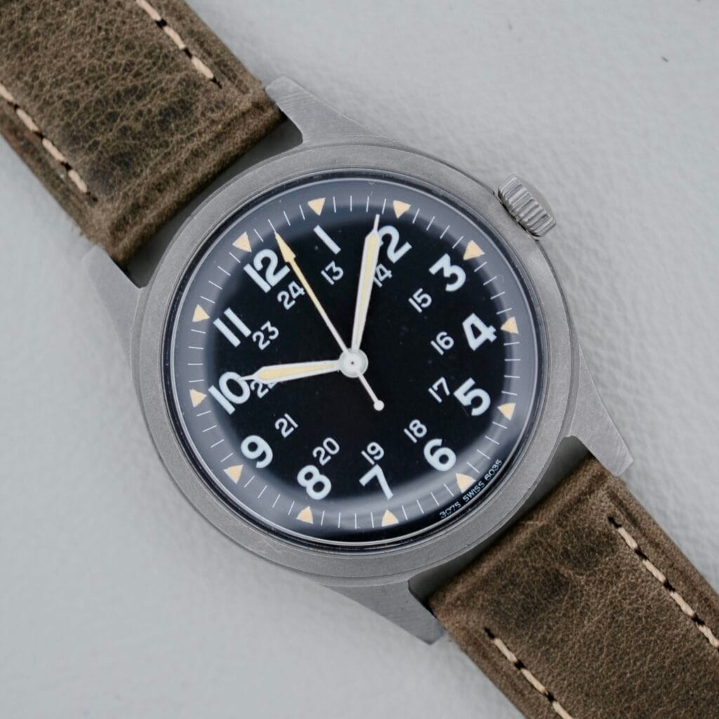 A 1974 Benrus GG-W-113 U.S. Military Issued watch with a black dial, white numerals, and brown leather strap is displayed on a light background—an iconic vintage military timepiece.