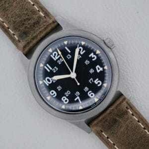 The 1974 Benrus GG-W-113 U.S. Military Issued wristwatch features a black dial with white numerals and hands, including a second hand, brown leather strap with visible stitching, and a brushed metal case against a light gray background.