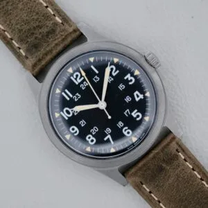 A 1974 Benrus GG-W-113 U.S. Military Issued watch with a black dial, white numerals, and brown leather strap is displayed on a light background—an iconic vintage military timepiece.