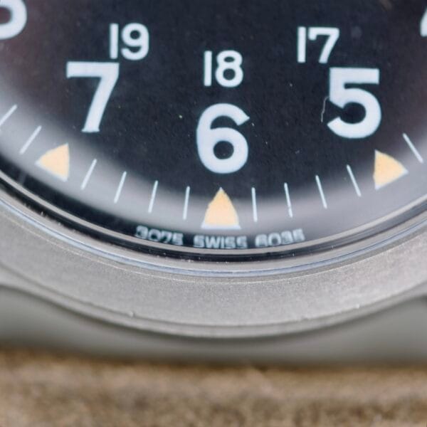 Close-up of the 1974 Benrus GG-W-113 U.S. Military Issued watch face, showing bold white numerals 6, 7, and 8 on a black background, SWISS MADE below the 6, and a metal casing.