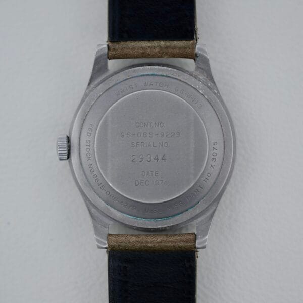 A close-up of the back of a 1974 Benrus GG-W-113 U.S. Military Issued wristwatch with a black leather strap. Its metal case shows serial numbers, manufacturing info, and DEC 1974 engraving as it rests on a light surface.