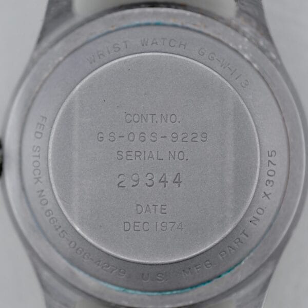 The back of a 1974 Benrus GG-W-113 U.S. Military Issued wristwatch shows engraved contract and serial numbers, date DEC 1974, and manufacturing details on its worn metal surface with visible scratches.