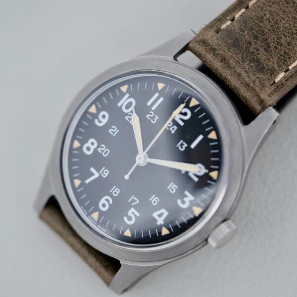 A close-up of the 1974 Benrus GG-W-113 U.S. Military Issued analog wristwatch featuring a black dial, white numerals, silver case, brown leather strap, and both 12-hour and 24-hour markings on the face.