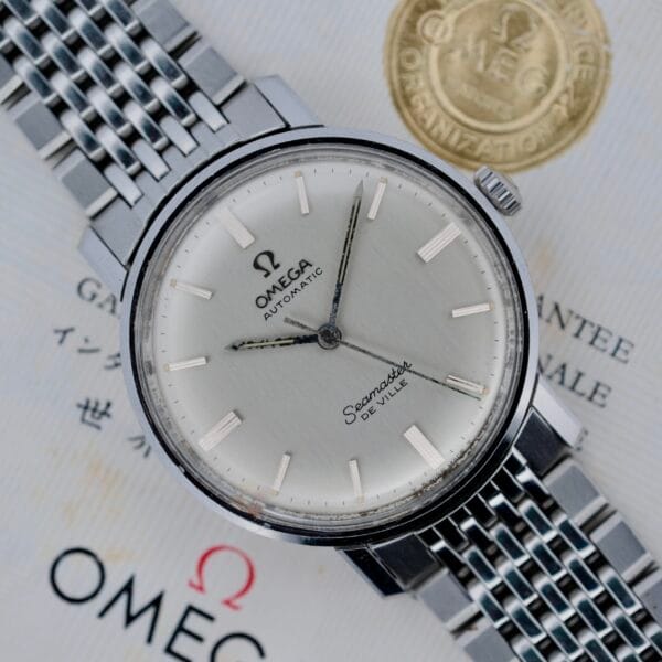 A close-up of a 1968 Omega Seamaster De Ville 165.020 automatic watch with a silver dial and metal bracelet, displayed atop official Omega documents bearing the brands logo and a gold seal.