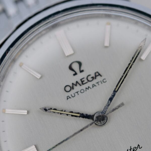 Close-up of a 1968 Omega Seamaster Deville 165.020 watch face displaying the Omega logo, hour markers, and aged hour and minute hands on a silver background.