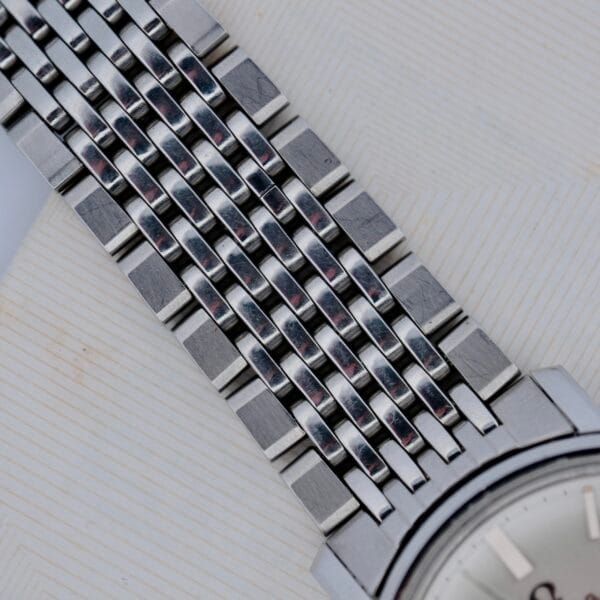 Close-up of a silver metal woven-link bracelet on a 1968 Omega Seamaster Deville 165.020, attached to a partially visible watch face, set against a textured off-white background.